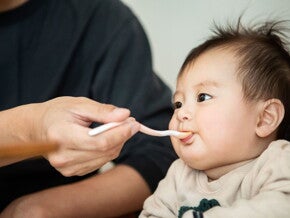 Bebé en silla alta, una mano adulta le alimenta papilla con una cuchara para bebés.