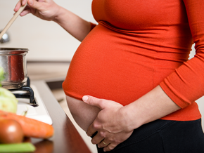 Mujer embarazada parada en frente de la cocina, preparando comida con ácidos grasos esenciales.