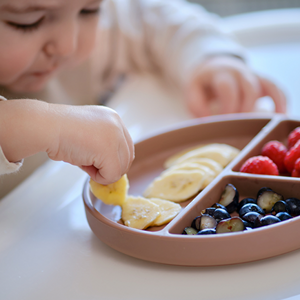 Alimentación saludable para bebés Un bebé de 8 a 12 meses comiendo con la mano un plato de frutas como bananas, frambuesas y uvas cortadas.