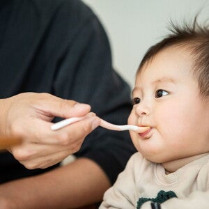 ¿Tu bebé está listo para los alimentos sólidos? Bebé en silla alta, una mano adulta le alimenta papilla con una cuchara para bebés.