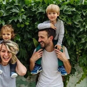 Crianza consciente y respetuosa Dos adultos con dos niños sobre sus hombros, frente a una pared cubierta de hojas
