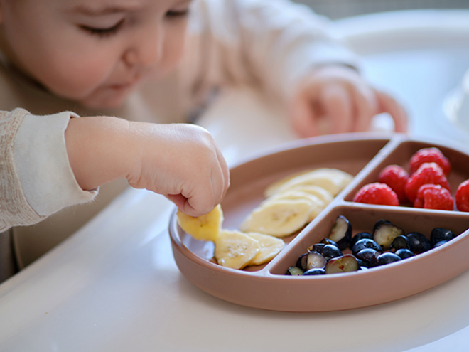 Un bebé de 8 a 12 meses comiendo con la mano un plato de frutas como bananas, frambuesas y uvas cortadas.