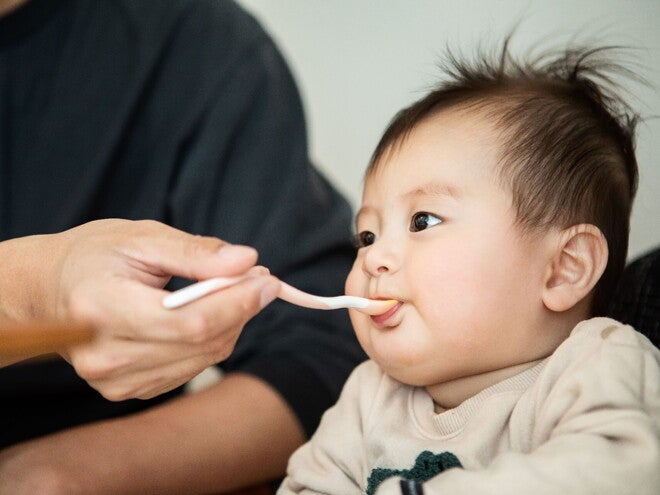 Bebé en silla alta, una mano adulta le alimenta papilla con una cuchara para bebés.