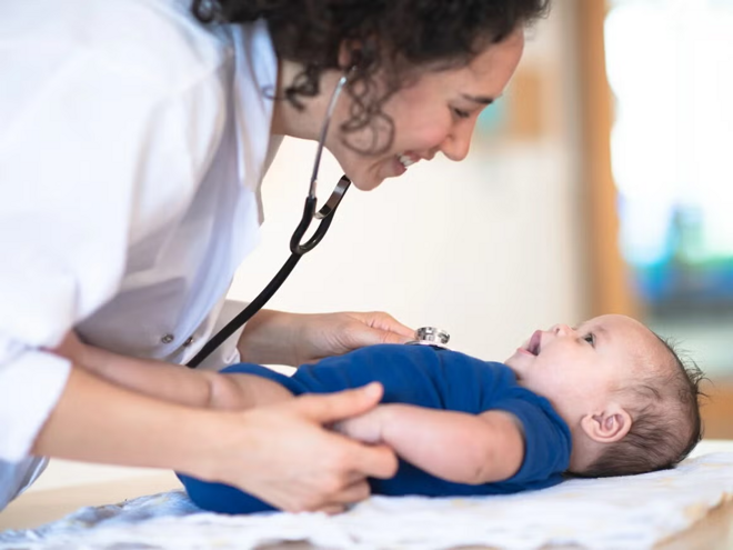 Bebé acostado en cama mientras la doctora le revisa y sonríe.