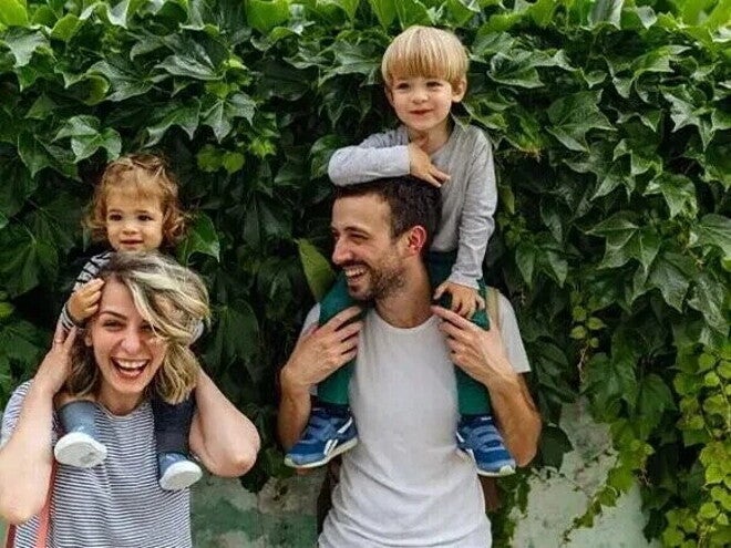 Crianza consciente y respetuosa Dos adultos con dos niños sobre sus hombros, frente a una pared cubierta de hojas