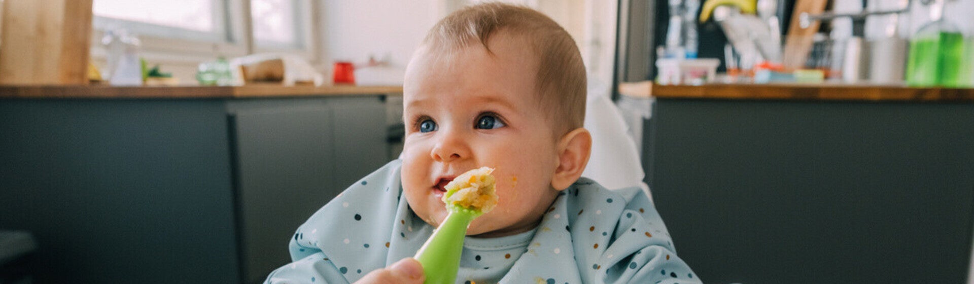 Mano de mamá dando de comer con una cuchara a su bebé que está sentado en silla alta. Alimentación complementaria del bebé.