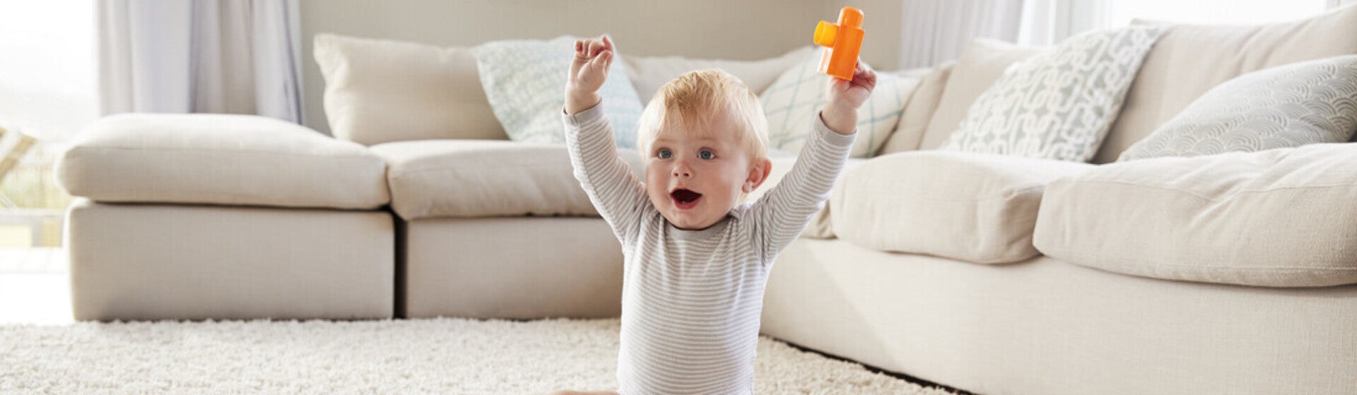 Bebé con juguetes sentando en la alfombra y levantando los brazos.