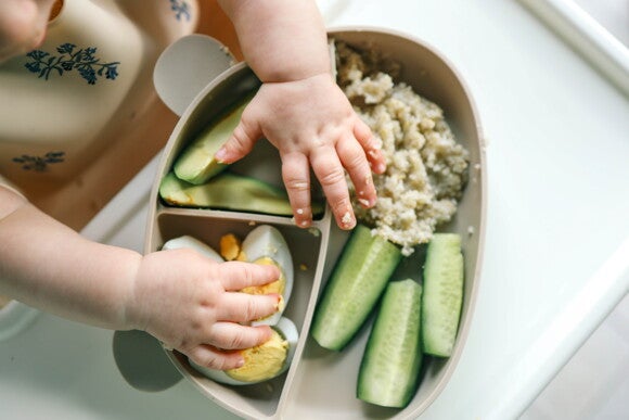Bebé en silla alta, manos de bebé tomando trozos de pepino y huevo, alimentos sólidos
